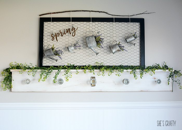 Black frame with chicken wire sits on top of a white shelf with metal watering cans hanging and a spring sign.
