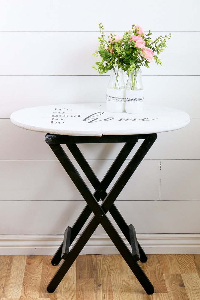 A small foldable bistro table has been painted black at the base and white on top with a saying it's so good to be home stenciled on the top. 