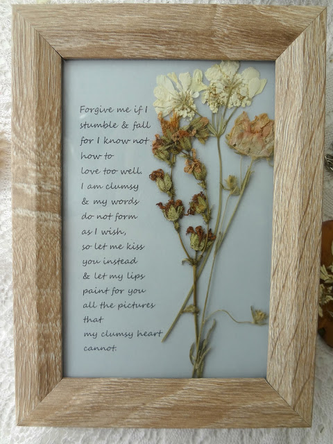 Spring flowers are dried and put inside of a wood frame with a saying written beside them. 