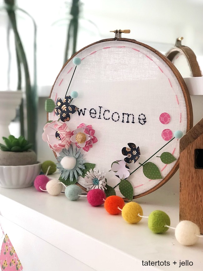 Make an embroidery hoop wall hanging. A hand-stitched word is surrounded by vibrant paper flowers in an embroidery hoop. Make one for your home or to give as a gift! 