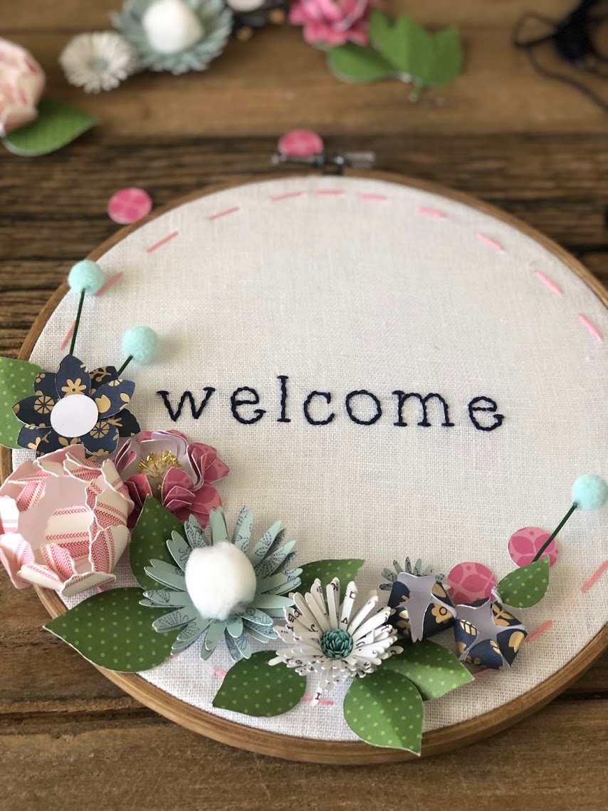 Make an embroidery hoop wall hanging. A hand-stitched word is surrounded by vibrant paper flowers in an embroidery hoop. Make one for your home or to give as a gift! 