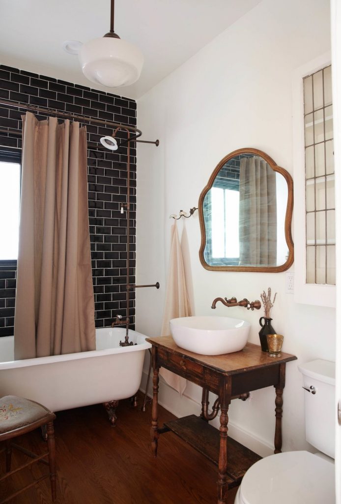 Masculine bathroom with white clawfoot tub, black subway tile and a vessel sink on an antique nightstand. 