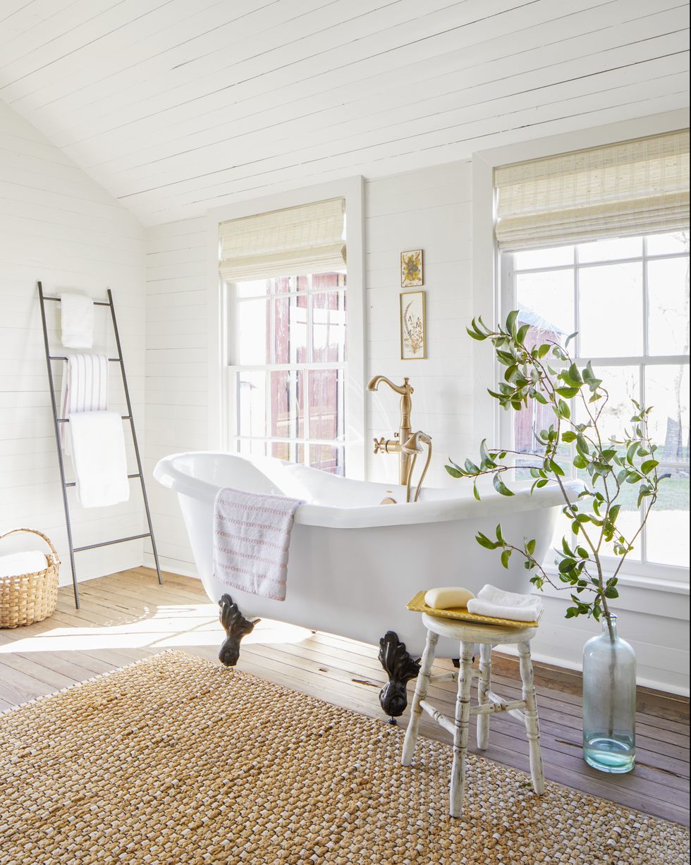 A white clawfoot tub with brass hardware against a shiplap white wall and windows. 