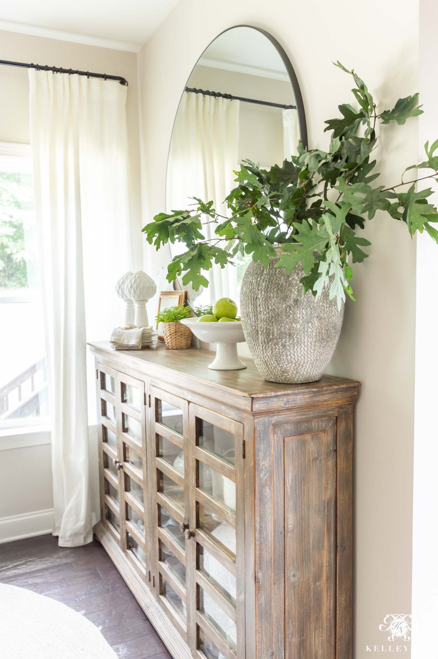 Green oak leaf branches in a tall chippy pottery vase on a dining room console with a footed vessel of green apples and napkins.