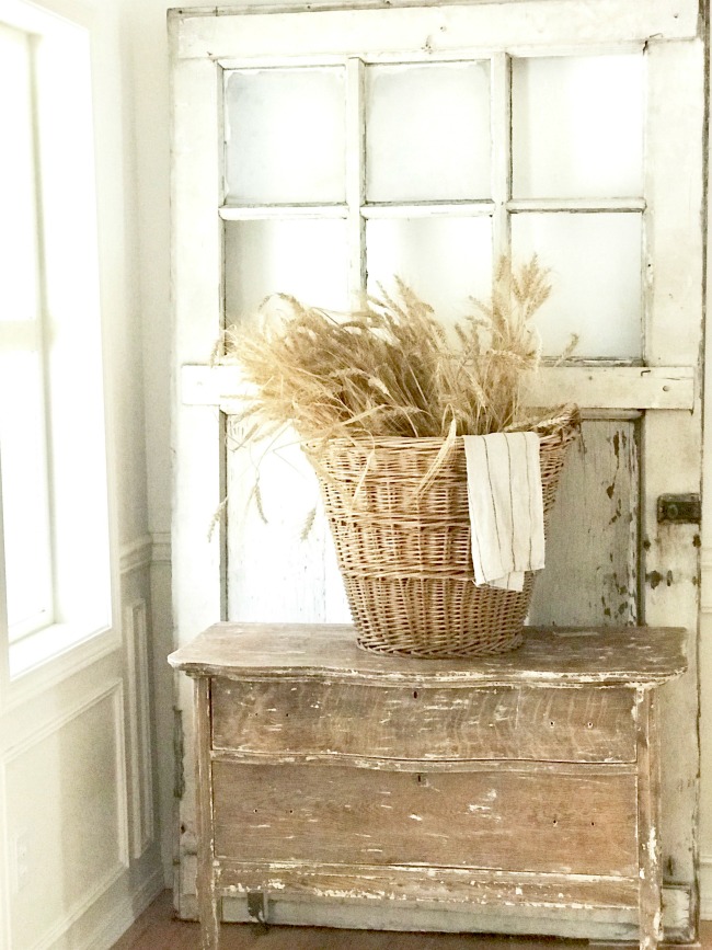 Natural wheat displayed in a vintage basket on a chippy.dresser and a vintage door behind. 