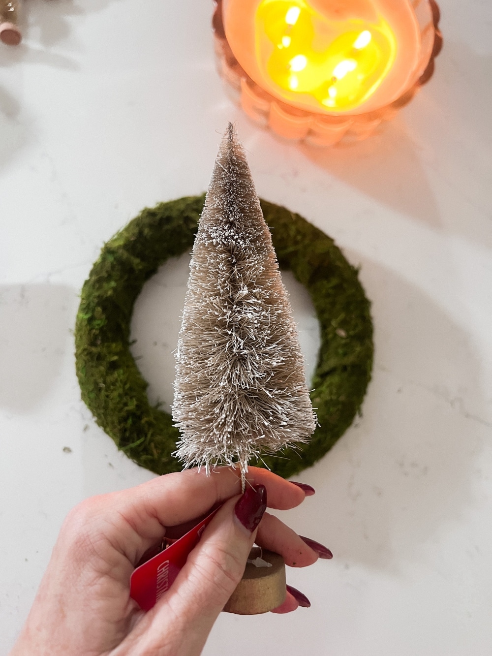 Cozy Dollar Store Christmas Centerpiece. Create a Terrain-inspired holiday centerpiece on a budget with dollar store finds! This DIY project combines bottle brush trees and a moss-covered wreath form, giving a high-end look without breaking the bank. Battery operated lights glow along with a candle in the center for a cozy addition to the holiday season.