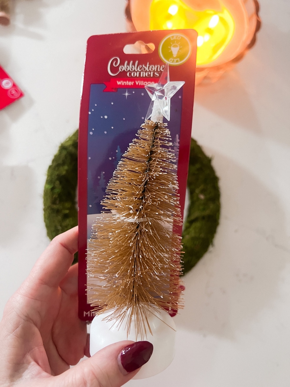 Cozy Dollar Store Christmas Centerpiece. Create a Terrain-inspired holiday centerpiece on a budget with dollar store finds! This DIY project combines bottle brush trees and a moss-covered wreath form, giving a high-end look without breaking the bank. Battery operated lights glow along with a candle in the center for a cozy addition to the holiday season.