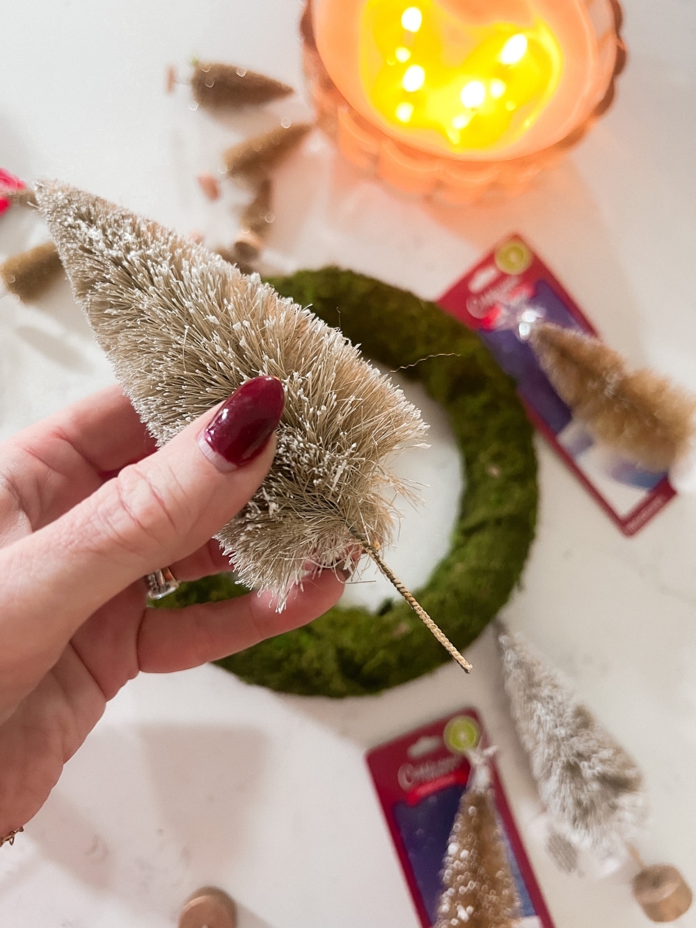 Cozy Dollar Store Christmas Centerpiece. Create a Terrain-inspired holiday centerpiece on a budget with dollar store finds! This DIY project combines bottle brush trees and a moss-covered wreath form, giving a high-end look without breaking the bank. Battery operated lights glow along with a candle in the center for a cozy addition to the holiday season.