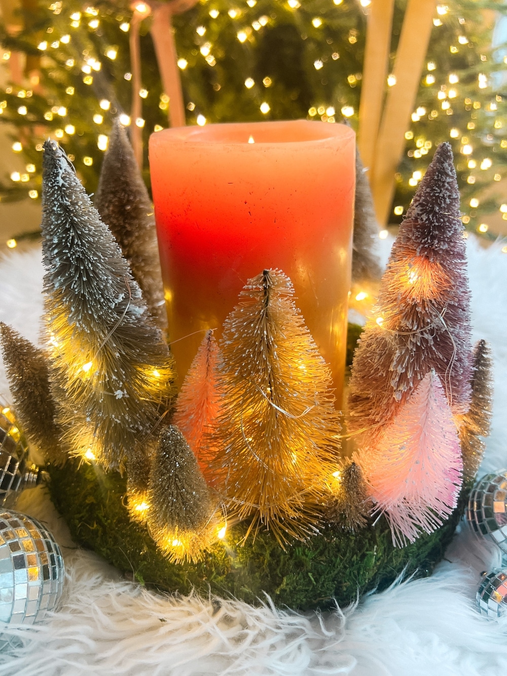 Cozy Dollar Store Christmas Centerpiece. Create a Terrain-inspired holiday centerpiece on a budget with dollar store finds! This DIY project combines bottle brush trees and a moss-covered wreath form, giving a high-end look without breaking the bank. Battery operated lights glow along with a candle in the center for a cozy addition to the holiday season.